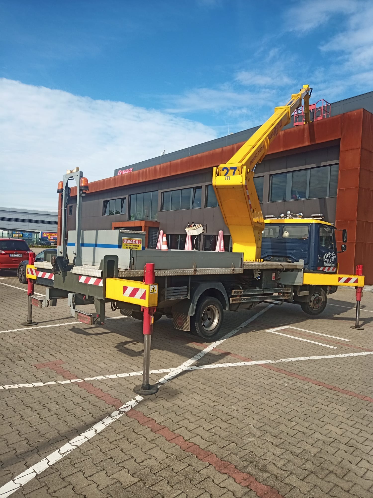 Żółto-niebieski podnośnik koszowy Mercedes ustawiony na parkingu przed budynkiem z brązową elewacją, z wysięgnikiem skierowanym w stronę dachu. Widoczne podpory stabilizujące pojazd i pomarańczowe...