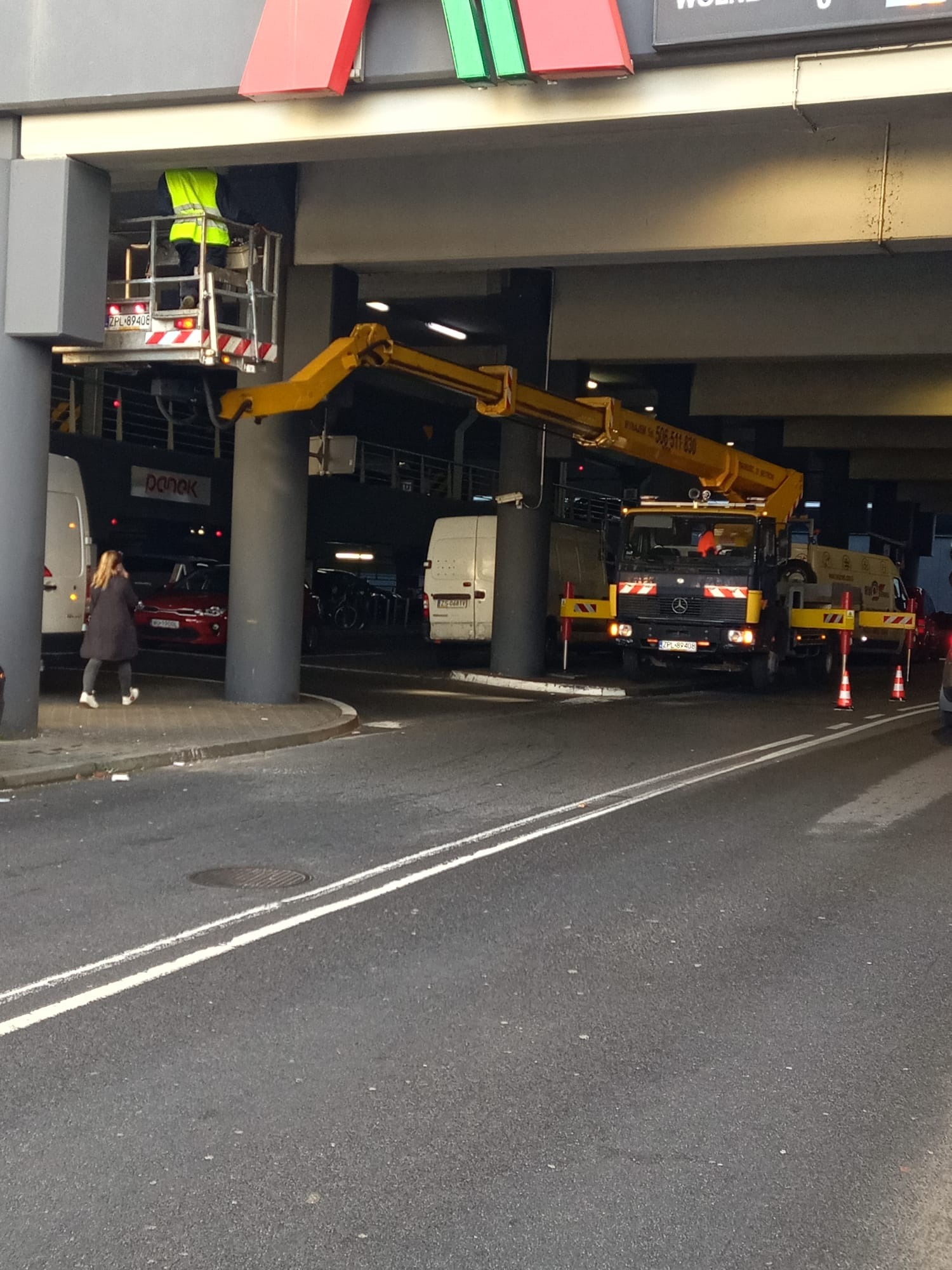Pracownik w żółtej kamizelce w koszu podnośnika na wysięgniku, prawdopodobnie naprawia lub czyści elementy konstrukcji pod zadaszeniem parkingu miejskiego, na pierwszym planie ulica z liniami,...