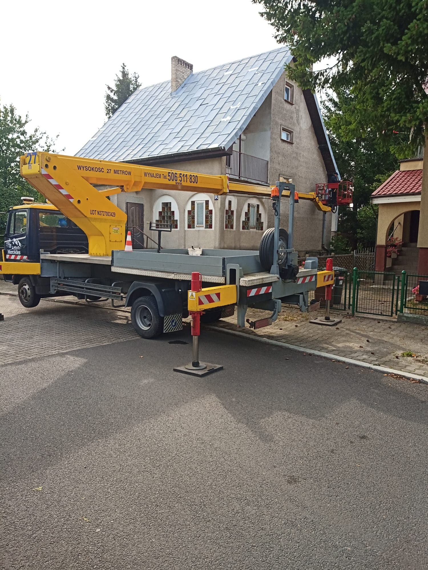 Żółty podnośnik koszowy Mercedes z wysięgnikiem ustawiony przed domem z szarą elewacją, osoba w koszu na wysokości okna, widoczny numer telefonu do wynajmu.
