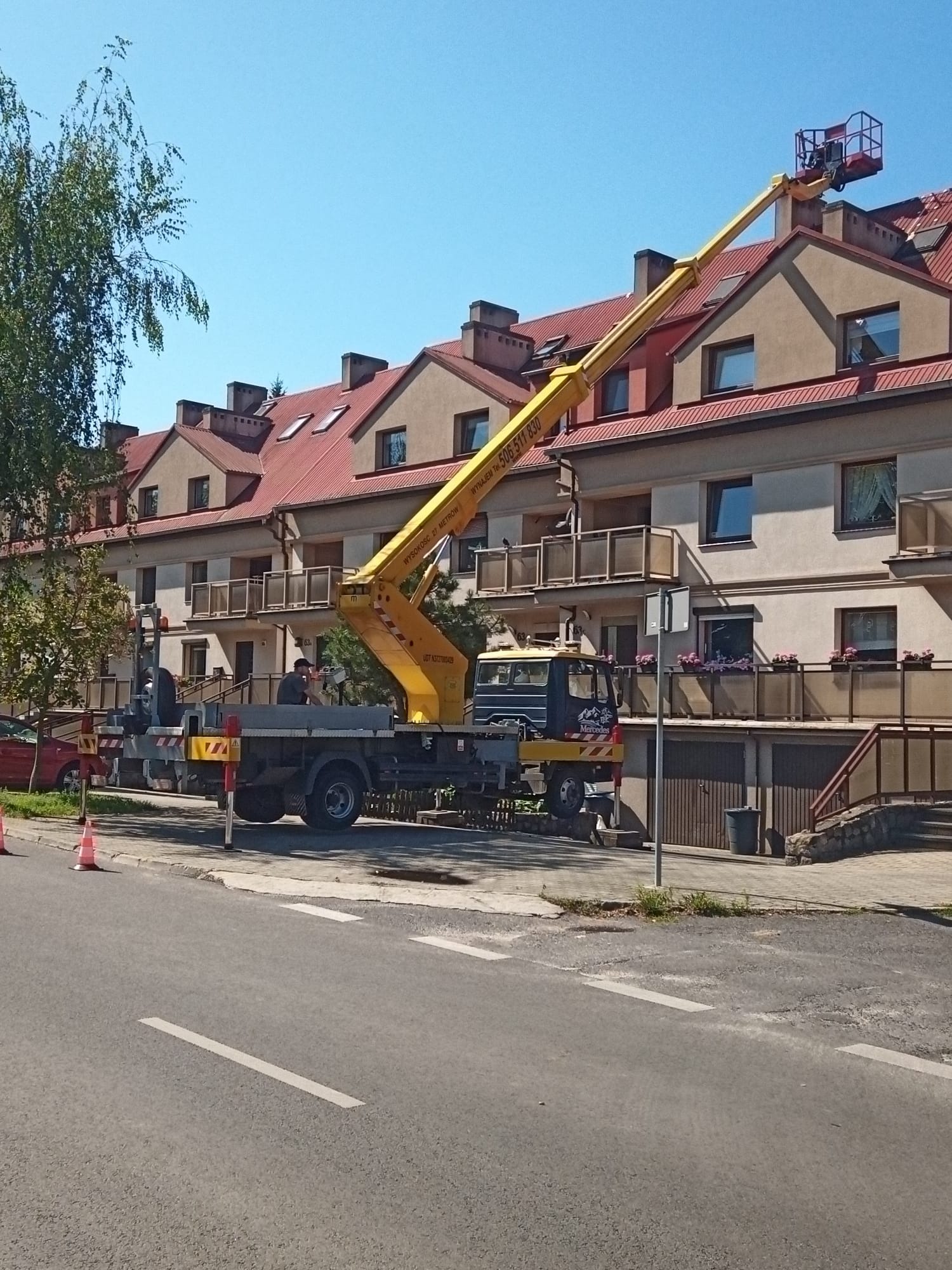 Żółty podnośnik koszowy na podwoziu ciężarówki Mercedes, zaparkowany na ulicy przed budynkiem mieszkalnym z czerwonym dachem, kosz podnośnika uniesiony w górę przy dachu budynku, widoczne barierki...