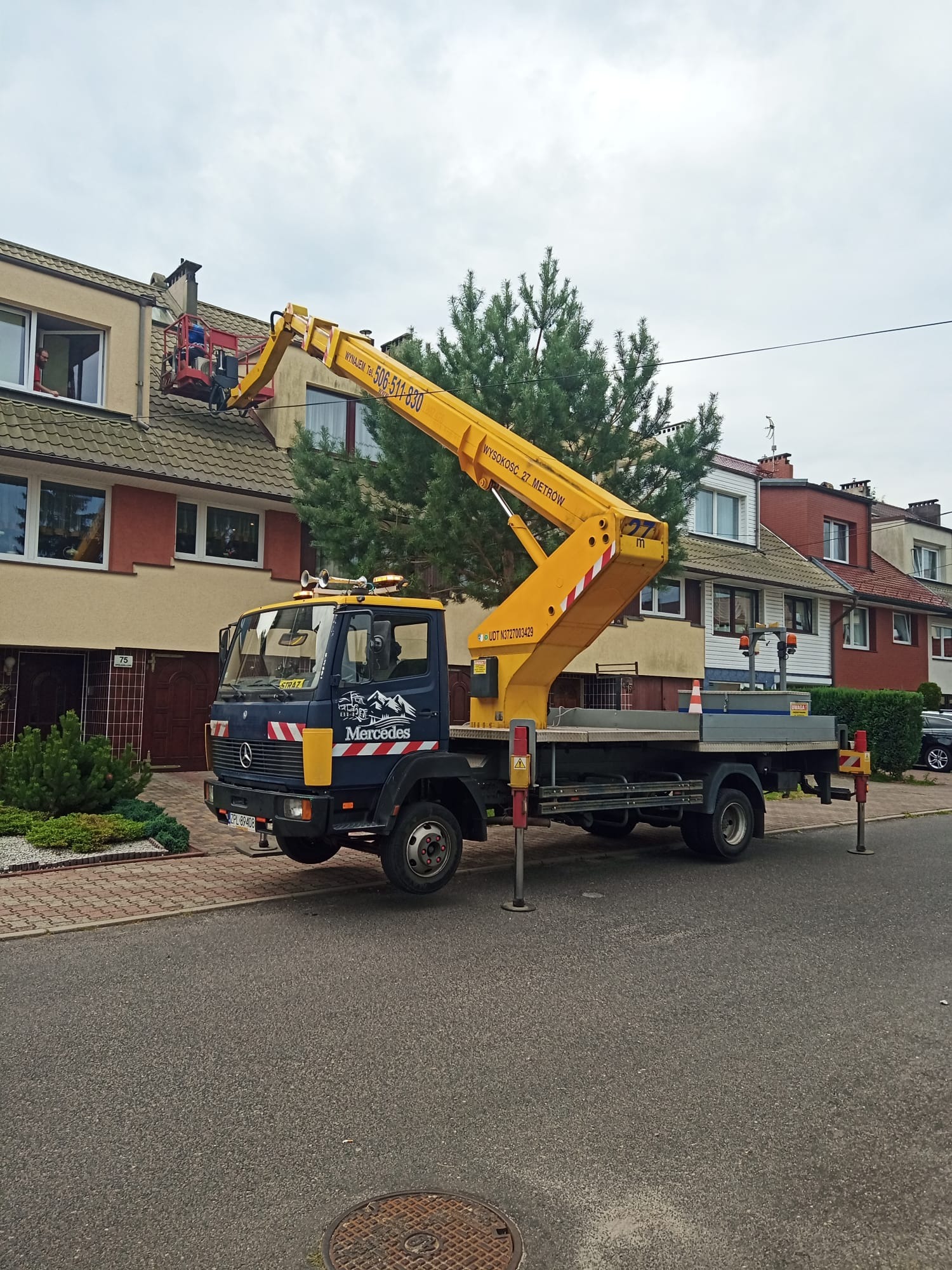 Żółty podnośnik koszowy Mercedes ustawiony na ulicy przed dwupiętrowym domem; pracownik w czerwonym koszu podnośnika pracuje przy kominie na dachu.