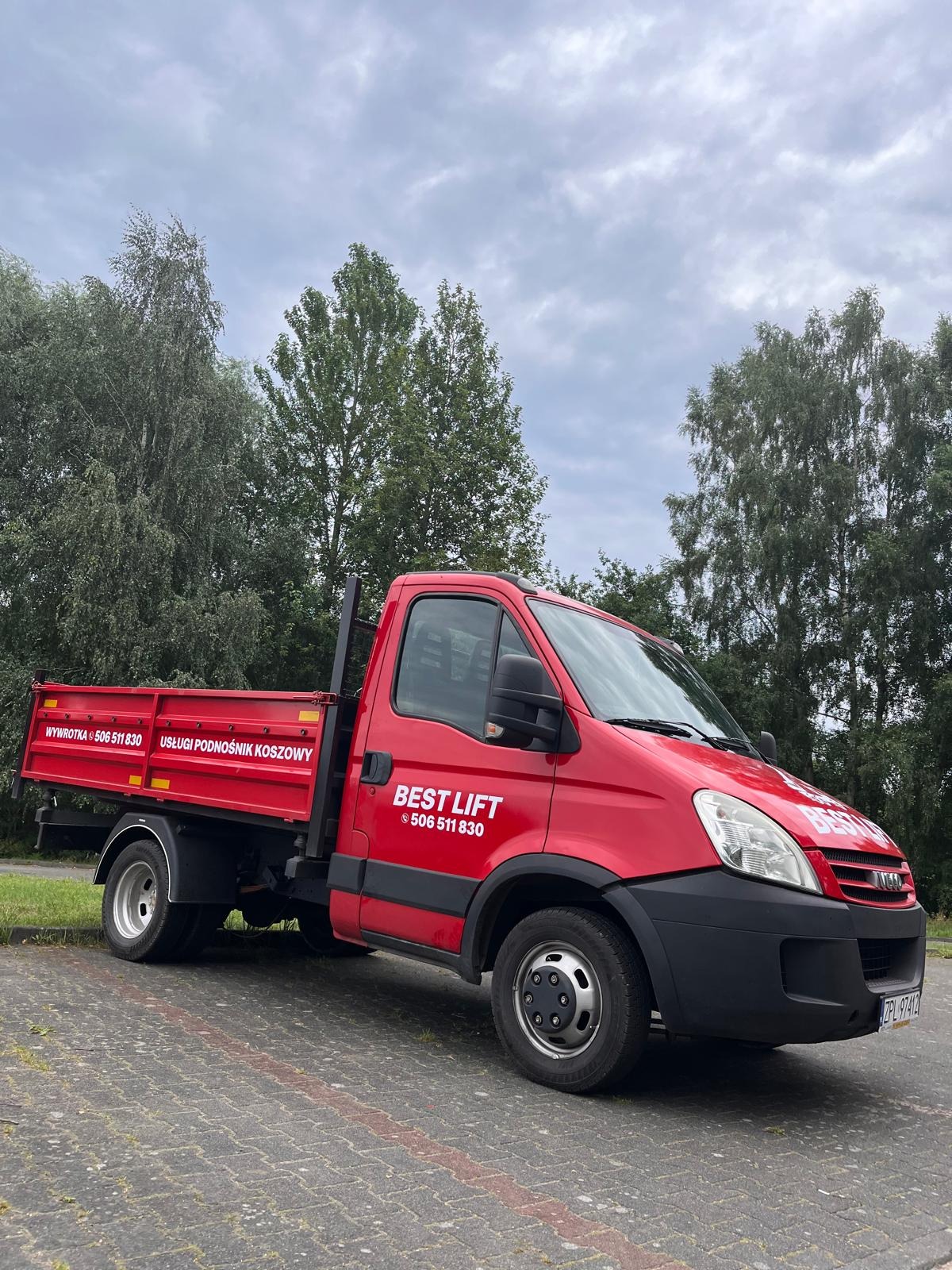 Czerwona ciężarówka z napisem 'BEST LIFT' i numerem telefonu na tle drzew i pochmurnego nieba, zaparkowana na kostce brukowej. Na skrzyni ładunkowej widoczny napis 'USŁUGI PODNOŚNIK KOSZOWY'.