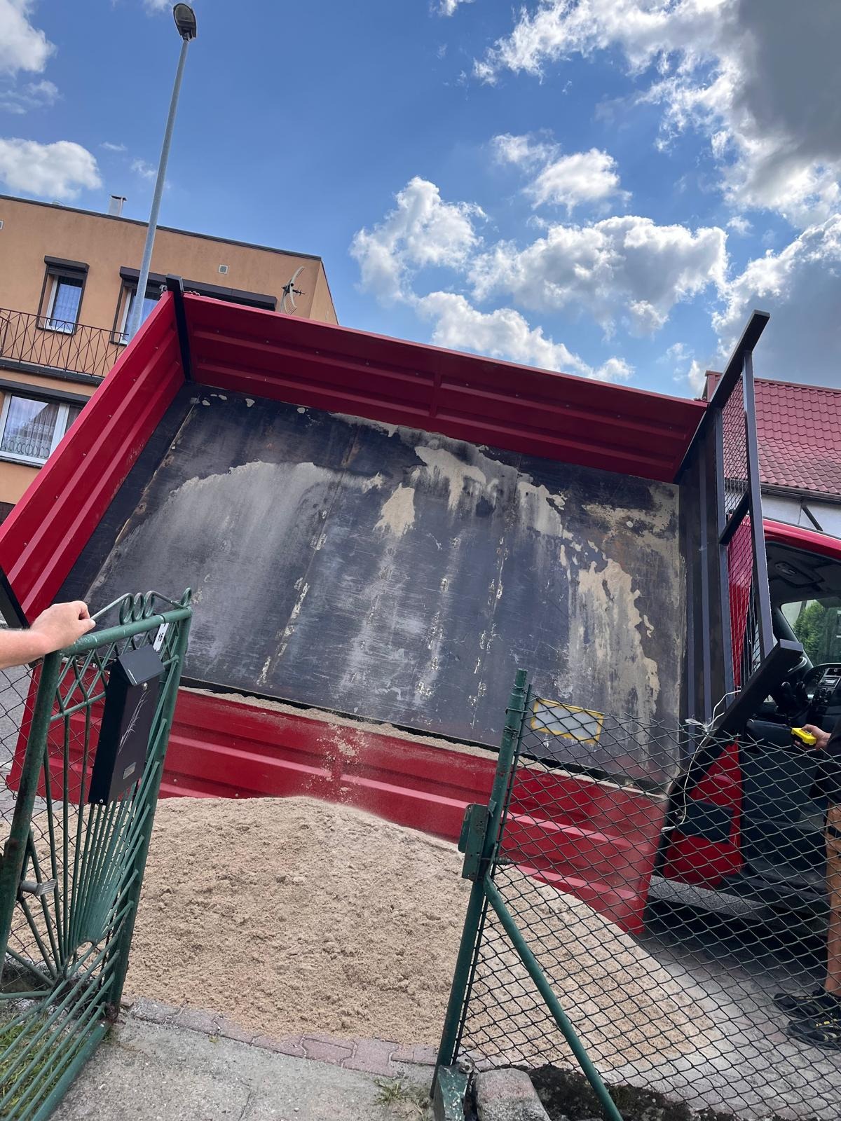Czerwona wywrotka z otwartą skrzynią ładunkową, wypełniona piaskiem, widziana zza zielonego ogrodzenia posesji, na tle budynku mieszkalnego i błękitnego nieba z chmurami.