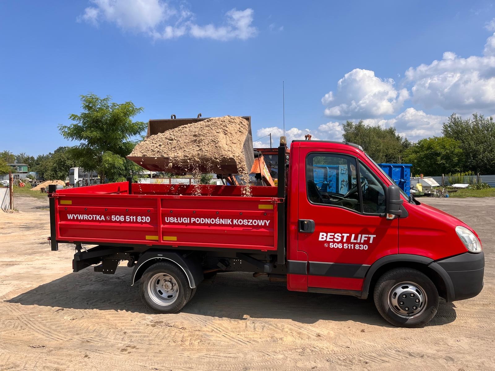 Czerwona wywrotka z podnośnikiem koszowym, z napisem 'BEST LIFT', wysypuje piasek na piaszczystym terenie w słoneczny dzień.