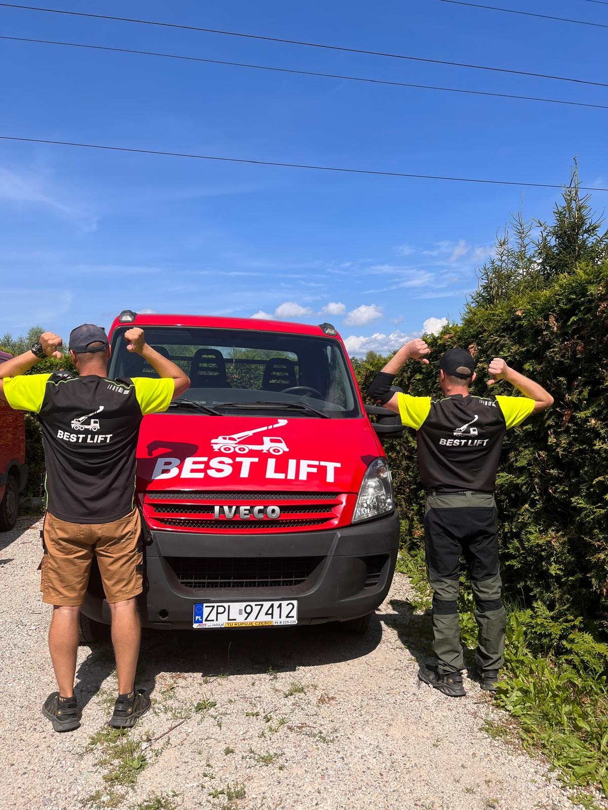 Dwóch pracowników firmy z Police, w koszulkach z logo firmy, prezentuje czerwony samochód dostawczy z logo firmy Best Lift na tle błękitnego nieba.