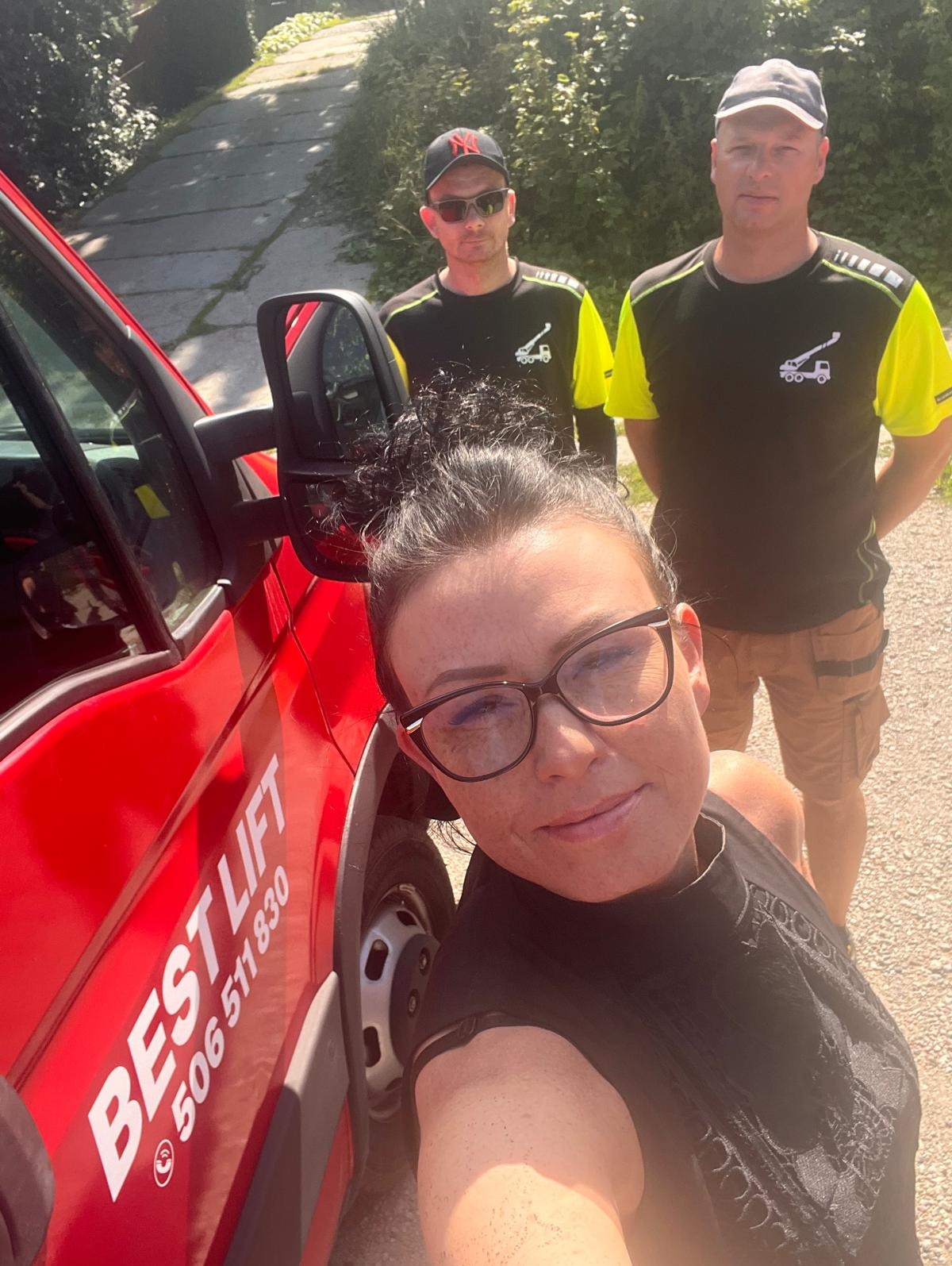 Autoportret kobiety w okularach na tle czerwonego samochodu z napisem 'BEST LIFT' i numerem telefonu, w tle dwóch mężczyzn w firmowych koszulkach z logo dźwigu
