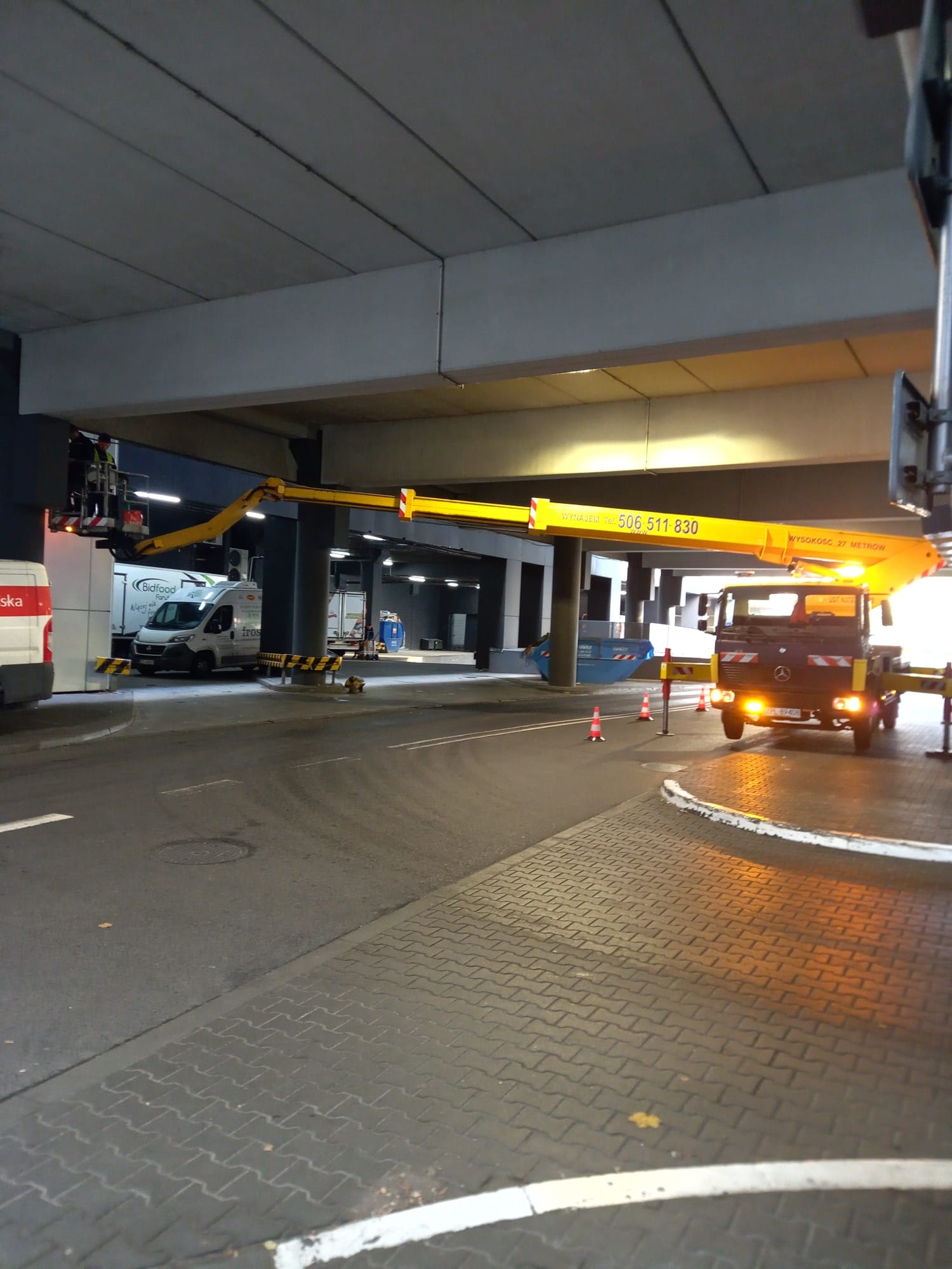 Żółty podnośnik koszowy z wysięgnikiem pod betonowym wiaduktem, pracownik w koszu, w tle zaparkowane samochody dostawcze.