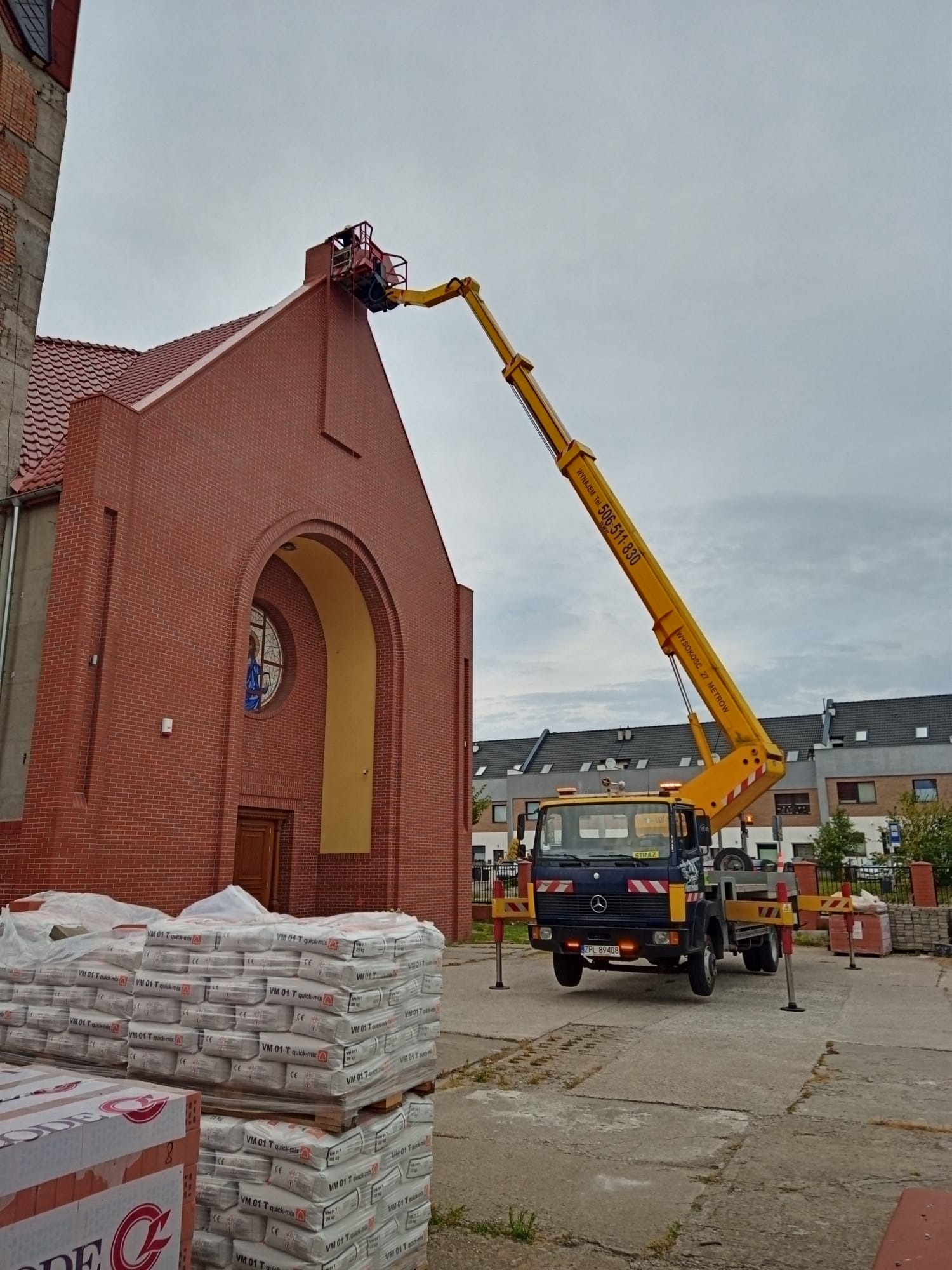 Żółty podnośnik koszowy przy ceglanym kościele z czerwoną dachówką, robotnik w koszu na wysokości, palety z zaprawą murarską na pierwszym planie, szare niebo w tle.