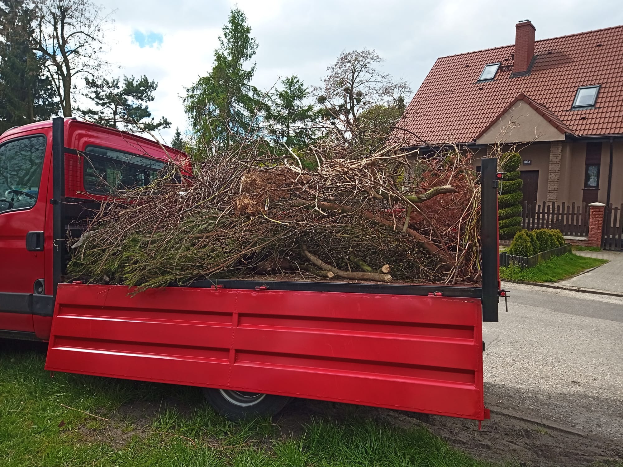 Czerwona ciężarówka z otwartą skrzynią załadowaną gałęziami i ściętymi drzewami na tle domu jednorodzinnego z czerwoną dachówką.