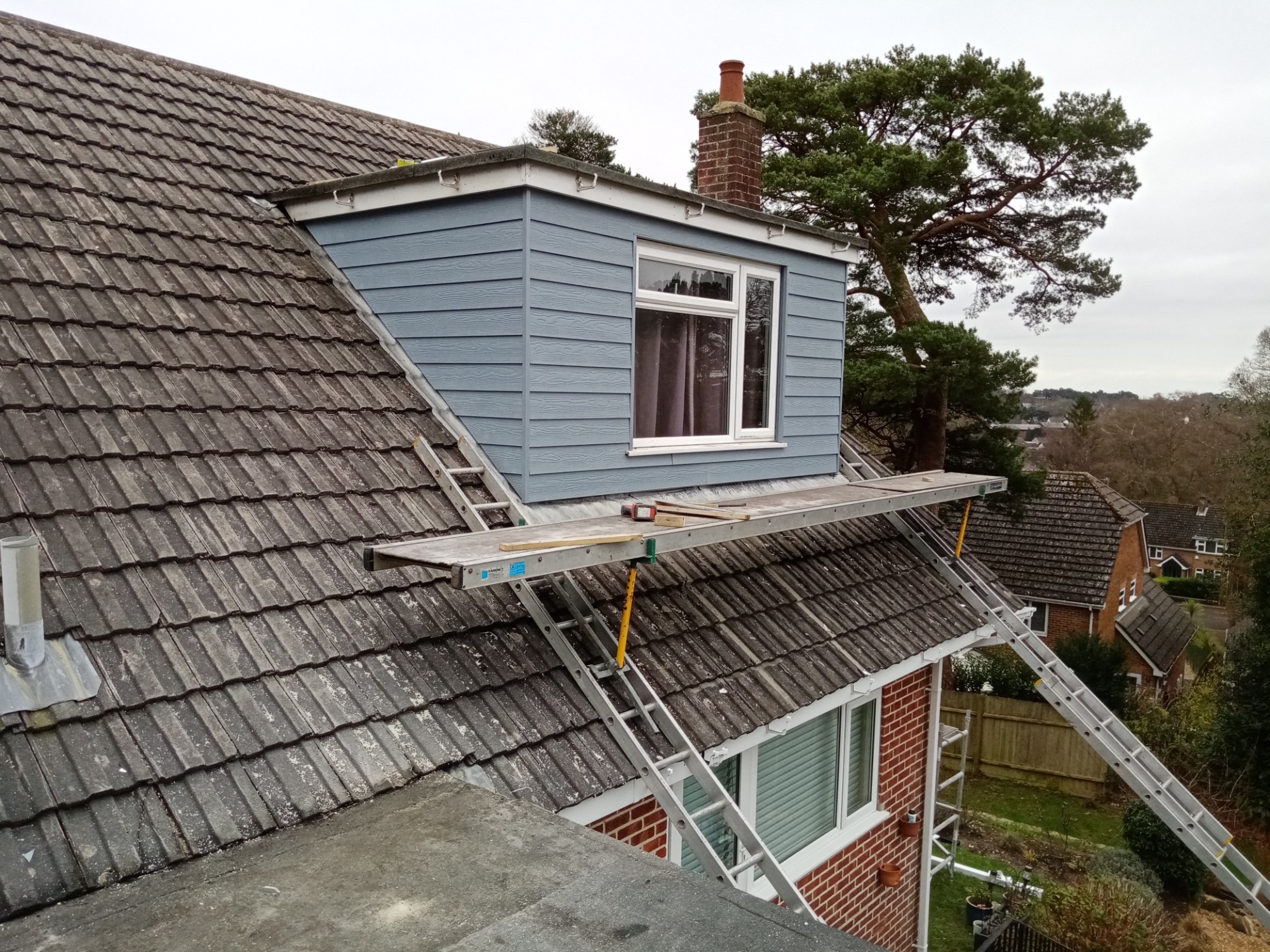 Niebieska nadbudówka na dachu z oknem, widoczna z rusztowania na dachu z dwoma drabinami opartymi o dom z cegły