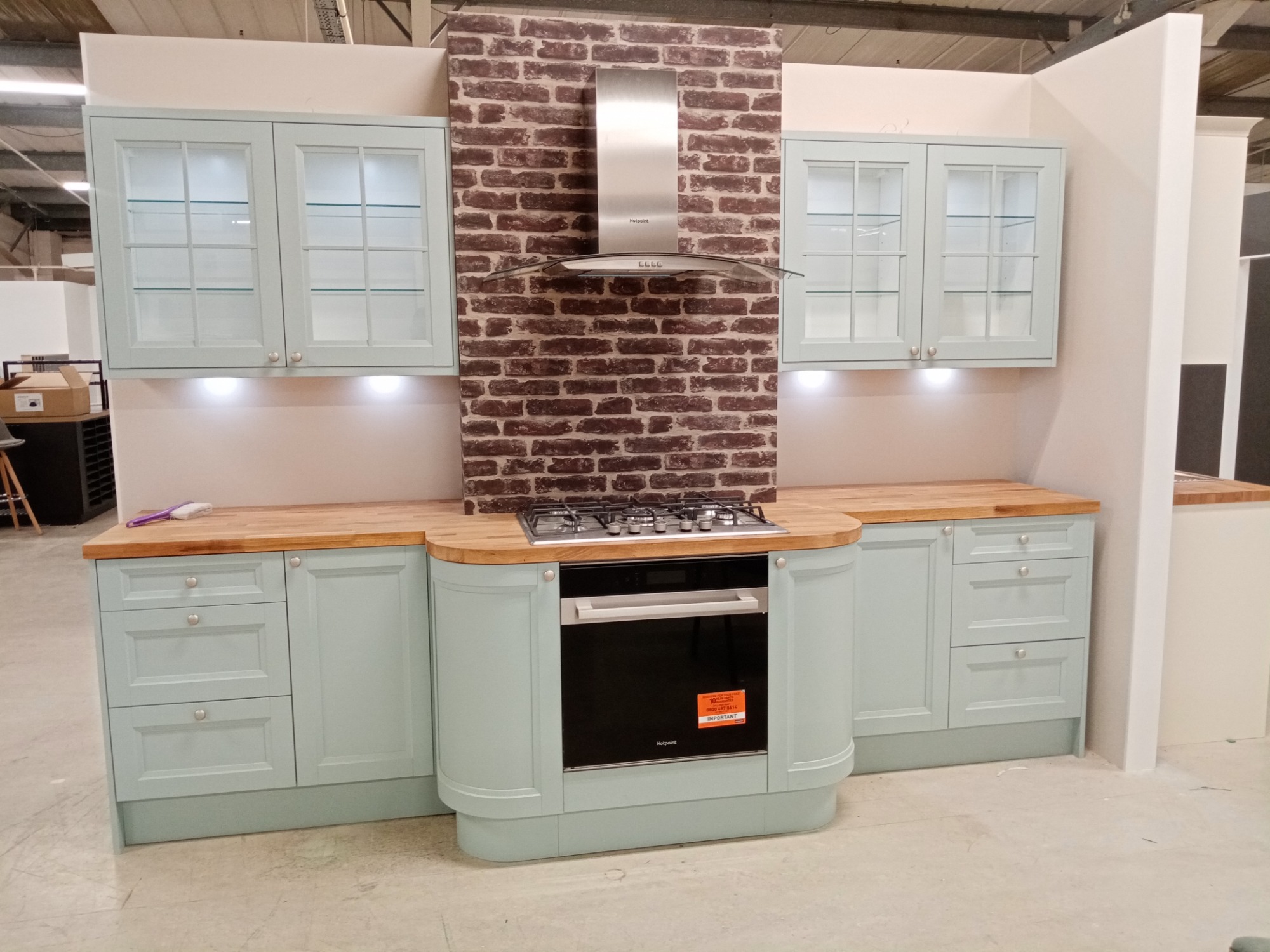 Jasnoniebieska zabudowa kuchenna z drewnianym blatem, piekarnikiem i płytą grzewczą na tle ściany z cegieł, prezentowana w sklepie.