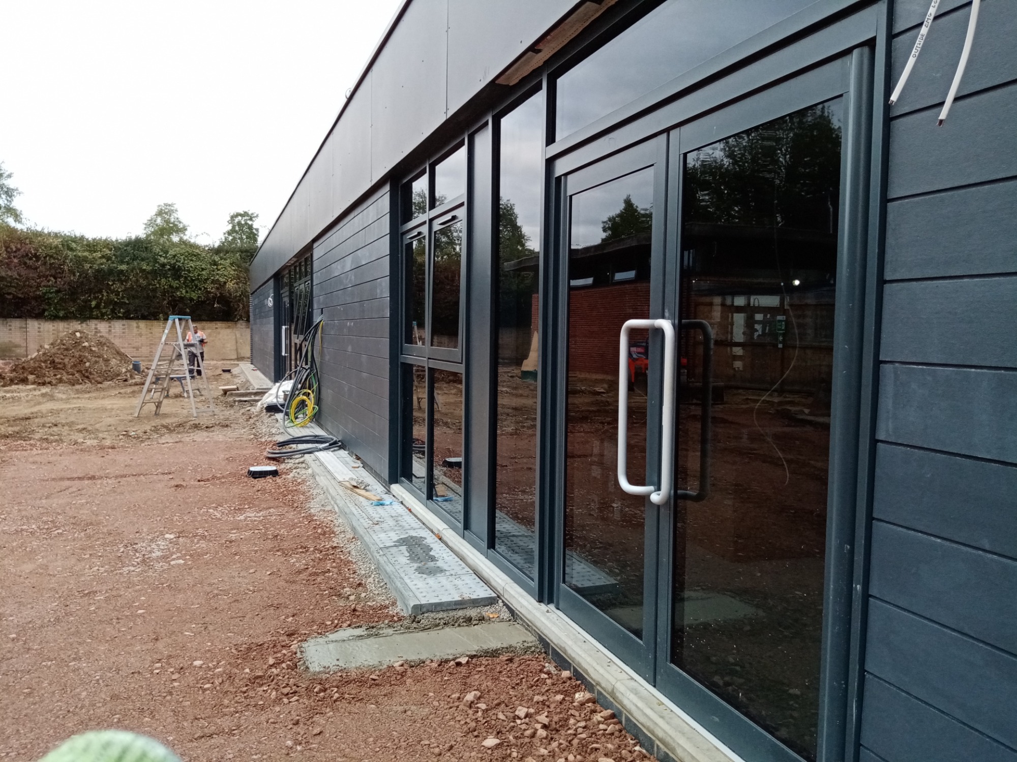 Montaż stolarki okiennej i drzwiowej w budynku z elewacją z paneli kompozytowych; widoczne kable i teren budowy.