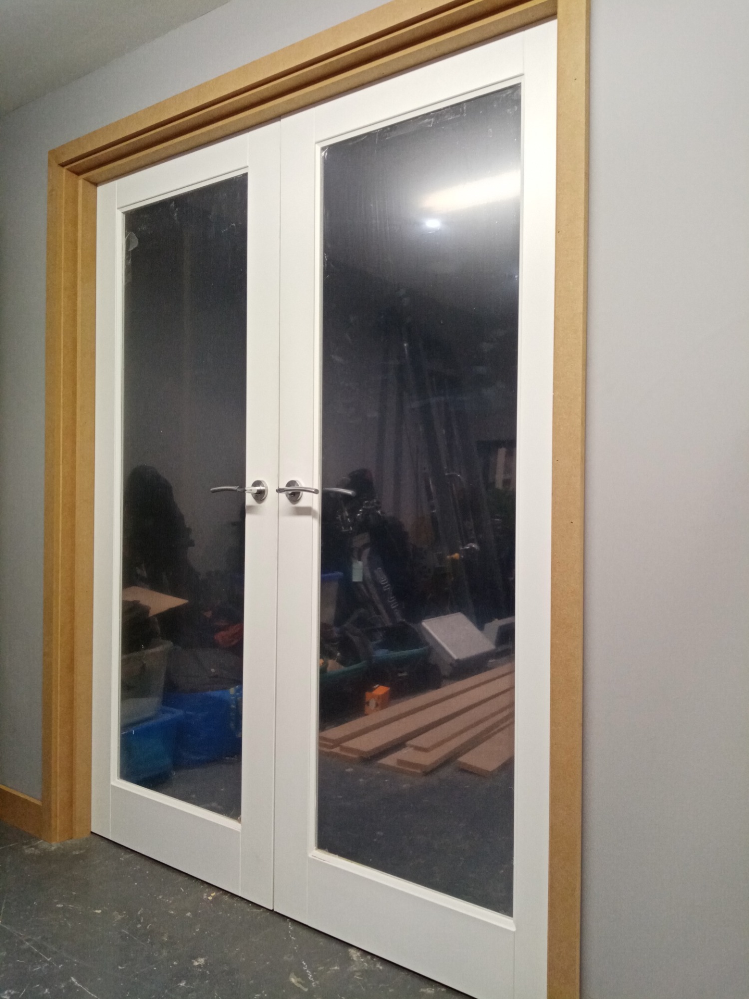 Podwójne, białe drzwi wewnętrzne z przeszkleniami i surową, drewnianą ościeżnicą, widok na zagracone wnętrze pomieszczenia.