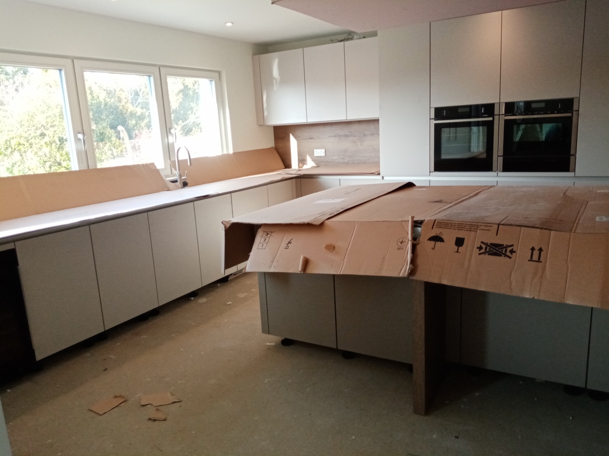 Jasna kuchnia z białymi szafkami i drewnianym blatem, częściowo zabezpieczona kartonem przed uszkodzeniem podczas prac wykończeniowych, widoczne dwa piekarniki w zabudowie.