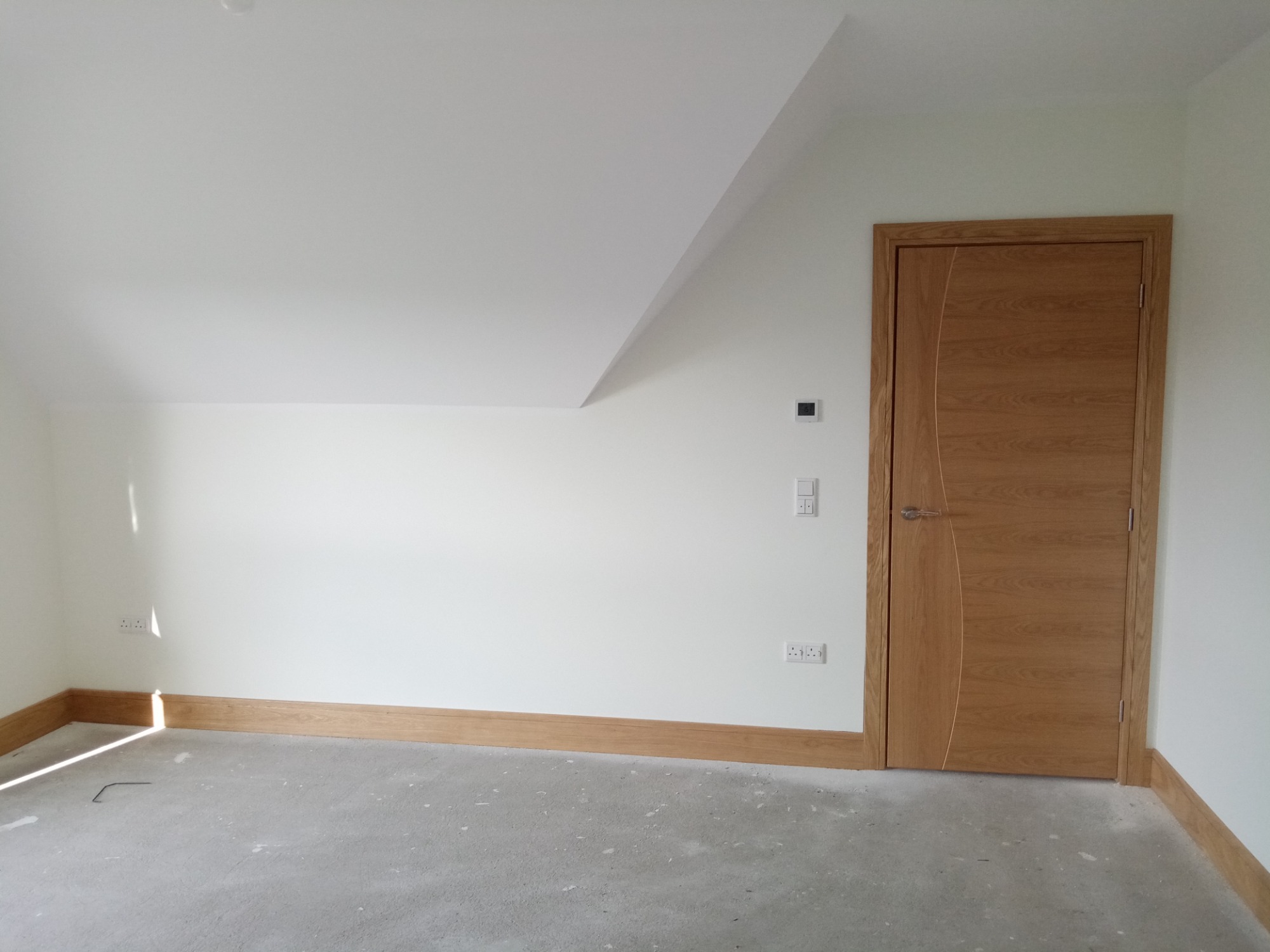 Pusta, jasna przestrzeń z nową, drewnianą ościeżnicą i drzwiami z widocznym rysunkiem drewna, listwy przypodłogowe również drewniane, surowa podłoga przed położeniem paneli lub płytek, instalacja...