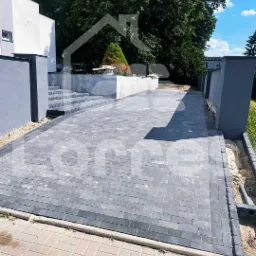 Szara kostka brukowa ułożona na podjeździe prowadzącym do domu z jasną elewacją, widoczne schody i elementy małej architektury ogrodowej.