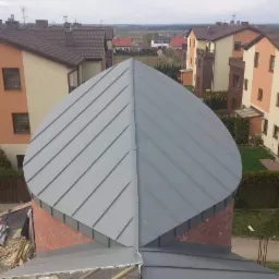 Kopuła z szarej blachy na ceglanej konstrukcji, widok z góry na osiedle domów jednorodzinnych w tle.