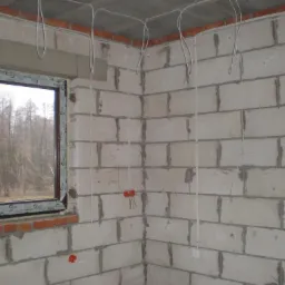 Surowy stan wnętrza z widoczną instalacją elektryczną: przewody zwisające ze stropu, puszki instalacyjne w ścianach z białego bloczka, okno z widokiem na drzewa.