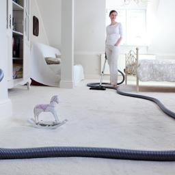 Daniel Urbański - Elektroinstalacje - Kobieta odkurza jasny dywan w pokoju z centralnym odkurzaczem, widoczny wąż podłączony do gniazdka w ścianie, w tle łóżko i białe meble, na pierwszym planie zabawkowy koń na biegunach.