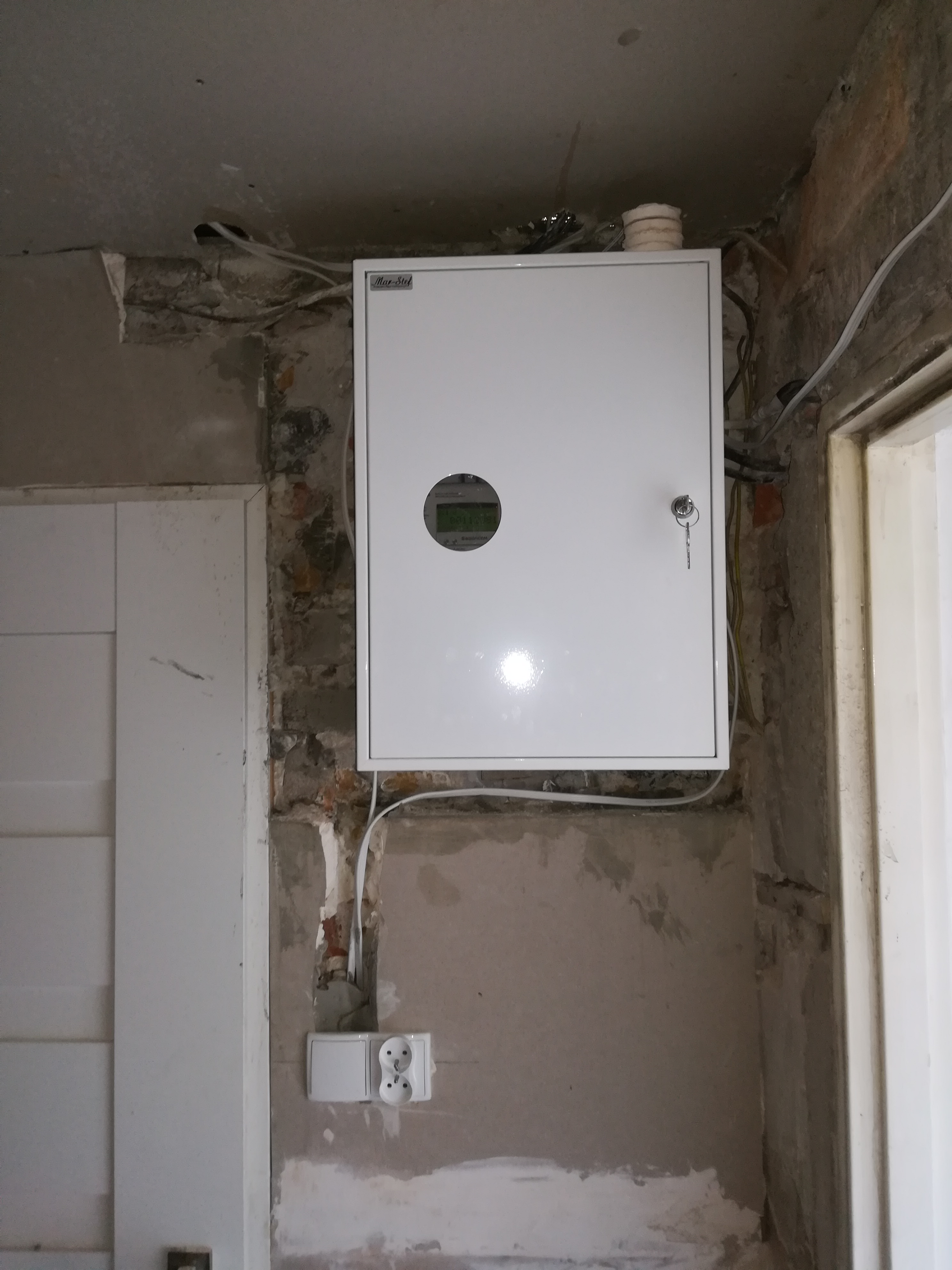 Biała skrzynka elektryczna z licznikiem, zamontowana na surowej ścianie w trakcie remontu, widoczne kable i gniazdko elektryczne.