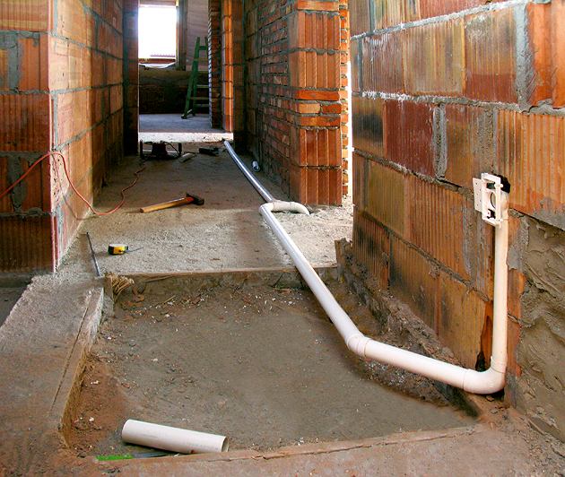 Wnętrze w trakcie prac instalacyjnych: biała rura PCV biegnie wzdłuż surowej ceglanej ściany, podłączona do puszki elektrycznej; widoczne narzędzia i fragment otwartego przewodu elektrycznego.