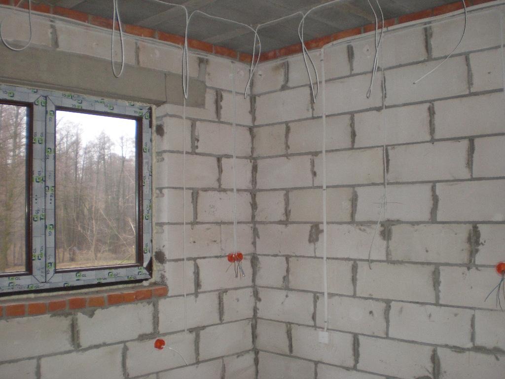 Surowy stan wnętrza z widoczną instalacją elektryczną: przewody zwisające ze stropu, puszki instalacyjne w ścianach z białego bloczka, okno z widokiem na drzewa.