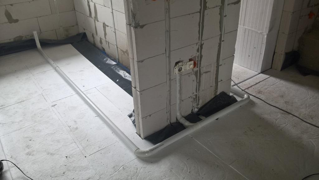 Instalacja elektryczna w trakcie montażu w surowym pomieszczeniu: białe rury prowadzone po podłodze i ścianach z białych bloczków, widoczne gniazdka elektryczne w ścianie, czarna folia izolacyjna...