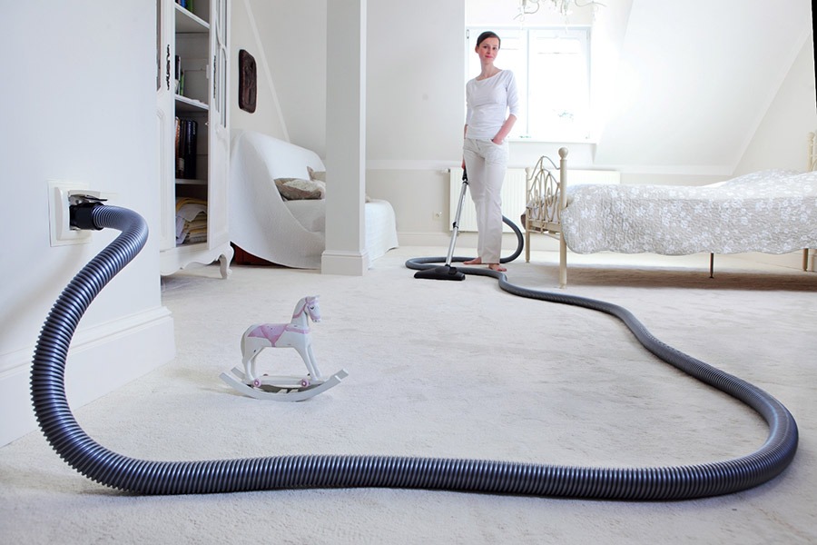 Kobieta odkurza jasny dywan w pokoju z centralnym odkurzaczem, widoczny wąż podłączony do gniazdka w ścianie, w tle łóżko i białe meble, na pierwszym planie zabawkowy koń na biegunach.