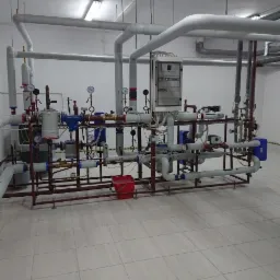 Skomplikowana instalacja grzewcza z licznymi rurami, zaworami, manometrami i sterownikiem elektronicznym, zamontowana w pomieszczeniu z białą podłogą i ścianami.