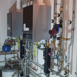 Dwa nowoczesne kotły gazowe Buderus zamontowane w piwnicy, widoczne liczne rury miedziane i stalowe, manometry oraz zbiornik wyrównawczy. Ściany piwnicy z widocznymi śladami wilgoci.