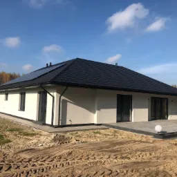 Nowoczesny parterowy dom z ciemnym dachem i panelami słonecznymi, widok z zewnątrz na częściowo utwardzony teren wokół budynku.