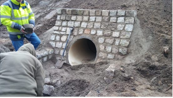 Budowa przepustu z kamienia w Mirsku: robotnicy układają kamienną obudowę wokół rury, widoczna łopata i zapas kamieni.