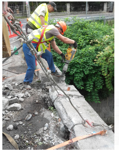 Dwóch pracowników w odblaskowych kamizelkach i kaskach, jeden z pomarańczowym kaskiem używa młota pneumatycznego do rozbiórki betonowej konstrukcji, która jest zabezpieczona stalowymi linami...