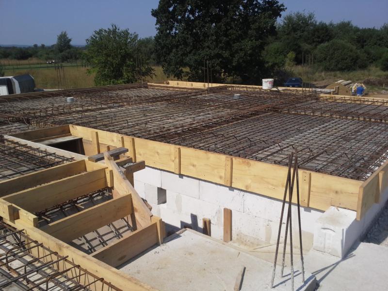 Zbrojenie stropu domu z bloczków, przygotowane do zalania betonem. Widoczne drewniane szalunki i siatka z prętów stalowych. W tle zieleń drzew i pola.