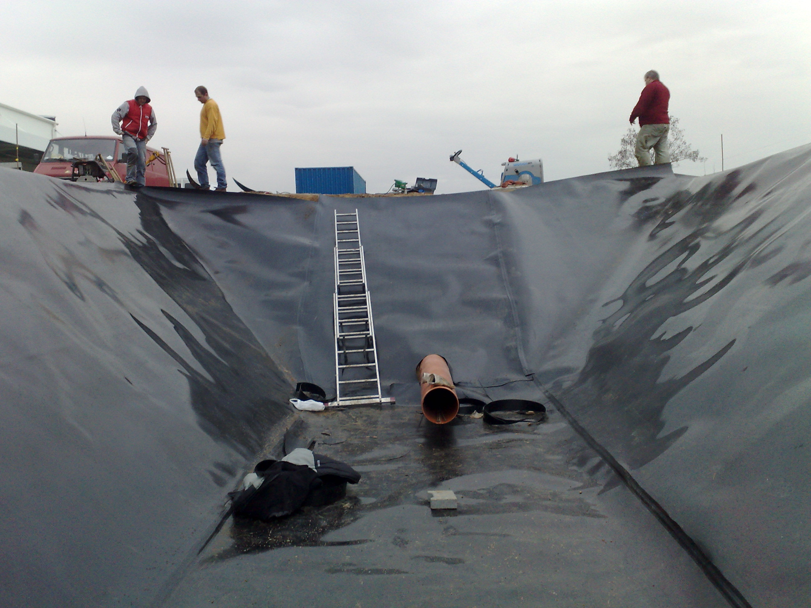 Montaż geomembrany w wykopie prawdopodobnie pod oczko wodne lub basen, widoczni pracownicy, drabina i sprzęt do zgrzewania folii.