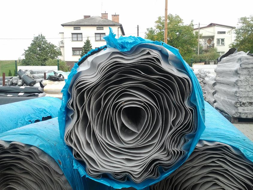 Rolki geomembrany wytłaczanej GEOKOMPOZYT TEFOND, zabezpieczone niebieską folią, składowane na zewnątrz, w tle budynki mieszkalne.