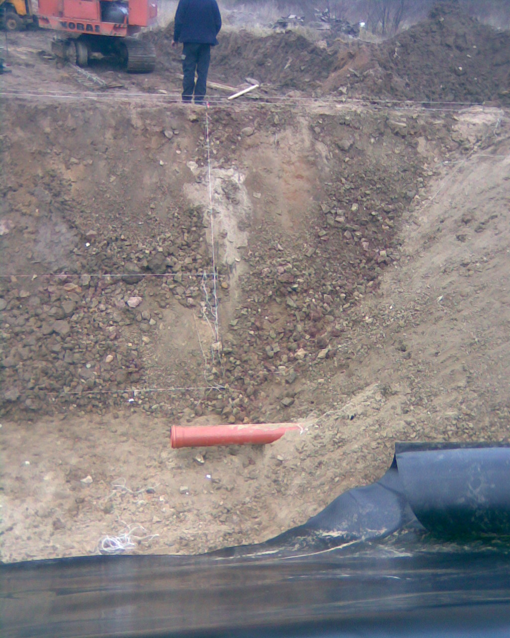 Wykop pod zbiornik wodny z widoczną rurą odpływową i częściowo rozłożoną geomembraną, na górze skarpy stoi osoba i koparka.