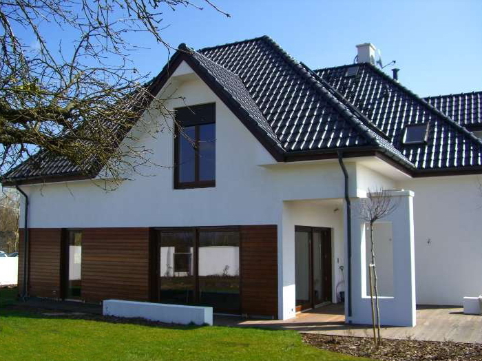 Nowoczesny dom jednorodzinny z białymi ścianami i ciemną dachówką, elementami drewnianej elewacji, widoczny z boku na tle zielonego trawnika i błękitnego nieba.