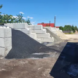 Skladowisko materiałów sypkich: czarny grys bazaltowy i jasny żwir, oddzielone betonowymi blokami w kształcie klocków lego, widoczny fragment naczepy samochodu ciężarowego.