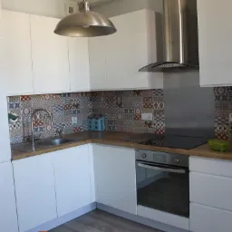 Nowoczesna kuchnia z białymi, lakierowanymi szafkami, drewnianym blatem, kolorowymi płytkami nad zlewem oraz stalowym okapem i lampą w stylu industrialnym.