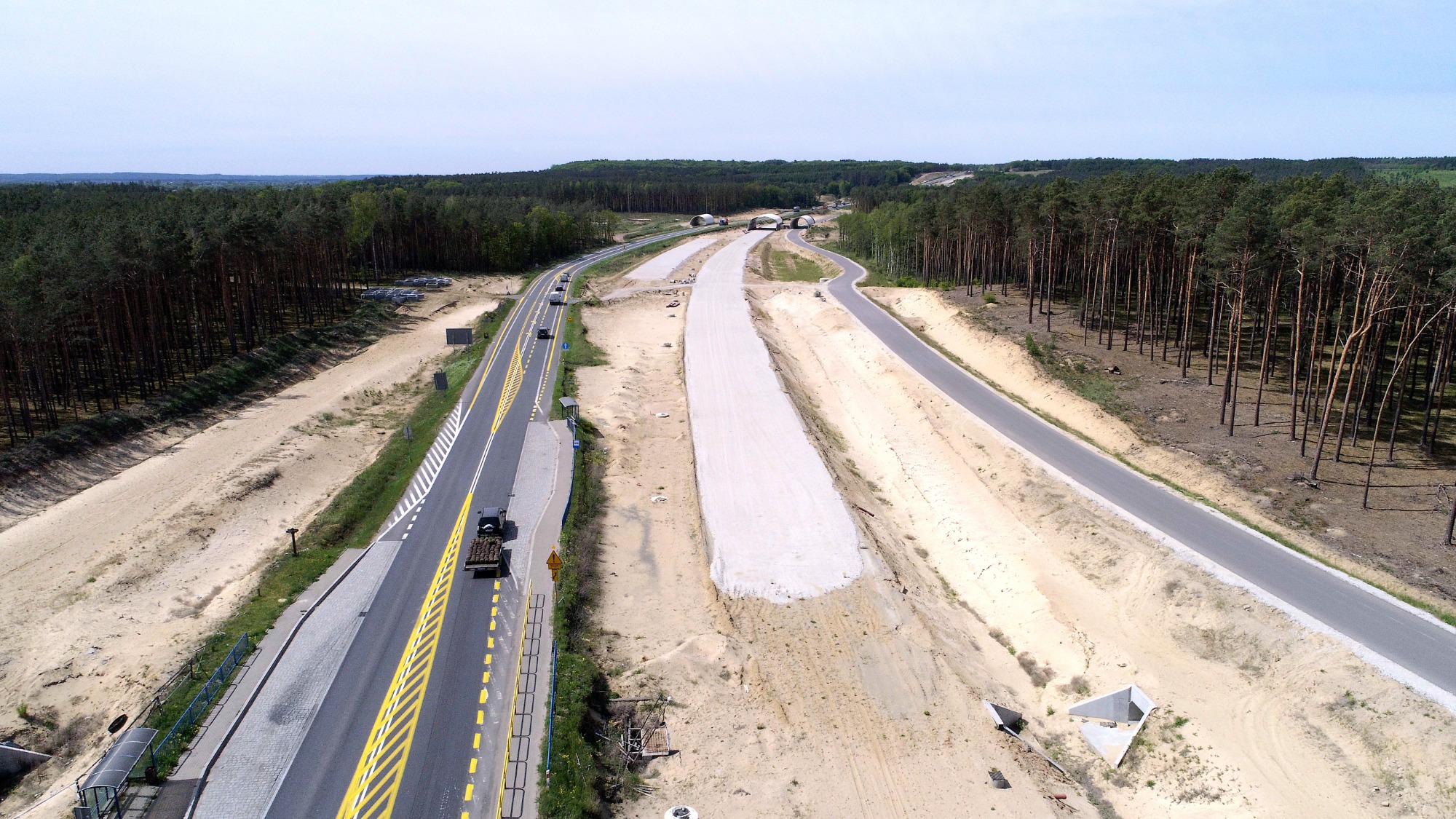 Zdjęcie z drona przedstawia budowę drogi ekspresowej z trzema pasami ruchu, widoczne tunele w oddali i lasy po obu stronach.