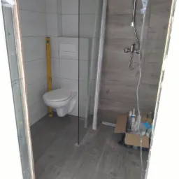 Wnętrze łazienki w trakcie wykańczania, widoczna podwieszana toaleta z żółtą poziomicą, częściowo zamontowana kabina prysznicowa i karton z materiałami budowlanymi.