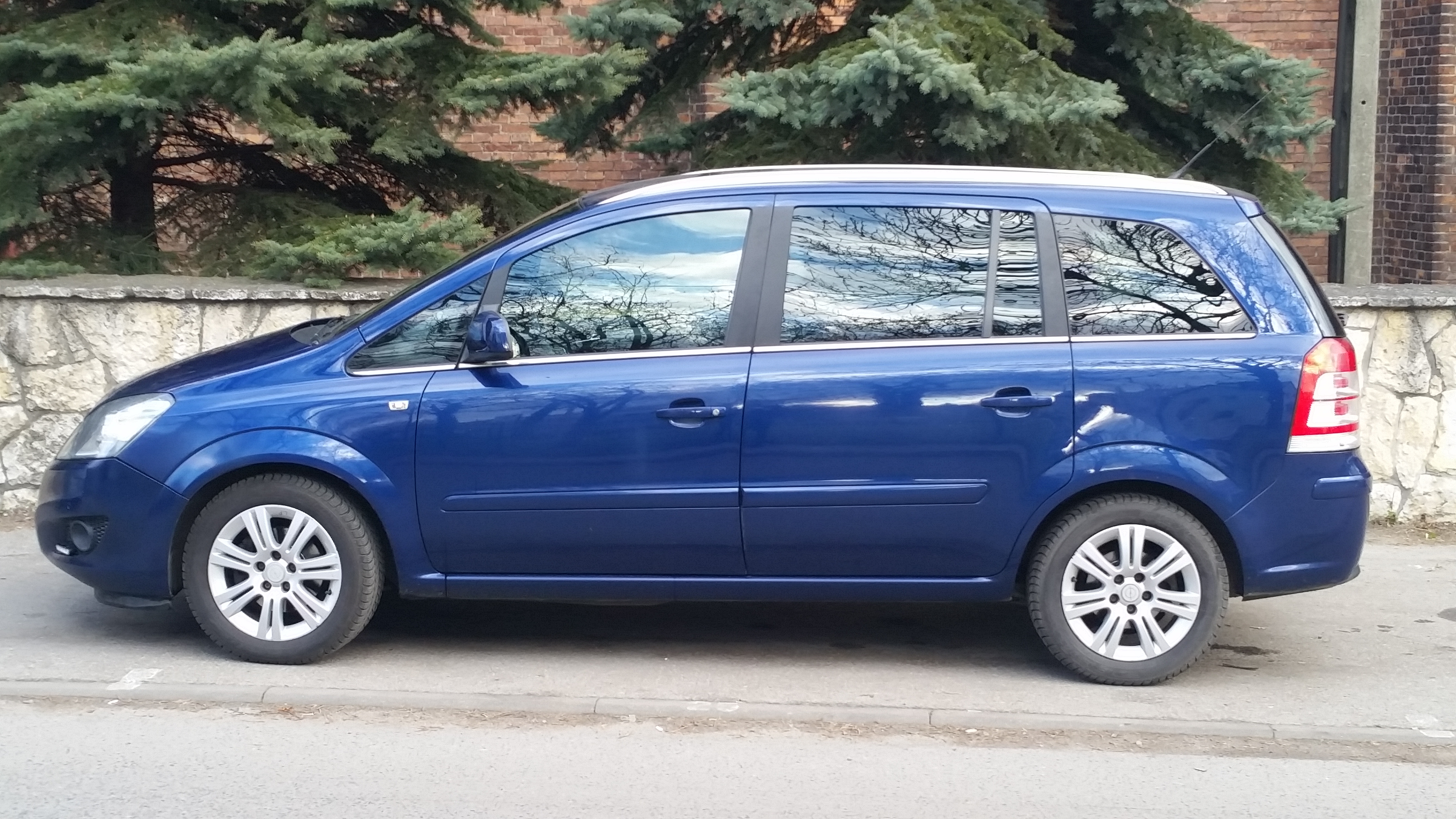 Niebieski minivan Opel Zafira zaparkowany na ulicy, odbicie drzew w oknach, widok z boku.