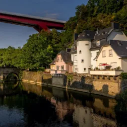 Malowniczy widok na rzekę Alzette w Luksemburgu, z kamiennymi budynkami o różnej architekturze na brzegu, odbijającymi się w wodzie, oraz czerwonym mostem w tle, wszystko w ciepłym świetle...