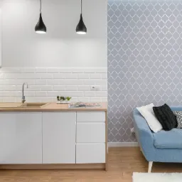 Nowoczesna kuchnia z białymi szafkami, drewnianym blatem i płytkami typu metro, połączona z salonem z niebieską sofą na tle ściany z geometrycznym wzorem.