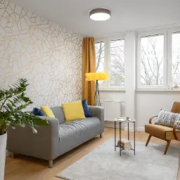 Jasny salon z geometryczną tapetą w odcieniach bieli i złota, szarą sofą z żółtymi i niebieskimi poduszkami, brązowym fotelem, dywanem w kolorze gołębim i wysoką rośliną w białej donicy.