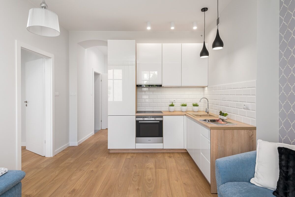 Jasna kuchnia w stylu skandynawskim z białymi błyszczącymi szafkami, drewnianym blatem, białymi płytkami metra i drewnianą podłogą; widok z salonu na otwartą kuchnię.