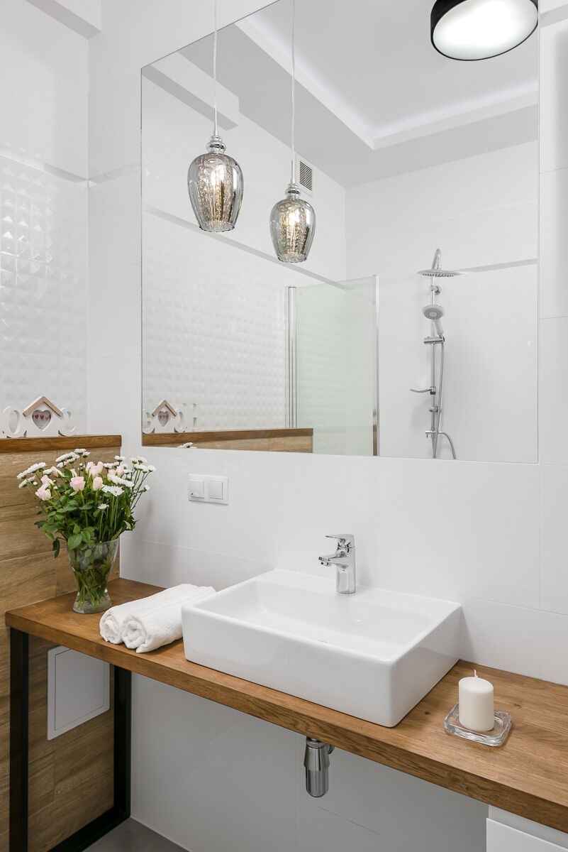 Nowoczesna łazienka z białą umywalką nablatową, drewnianym blatem, lustrem, szklanymi lampami wiszącymi i dekoracjami: kwiatami oraz świecą.