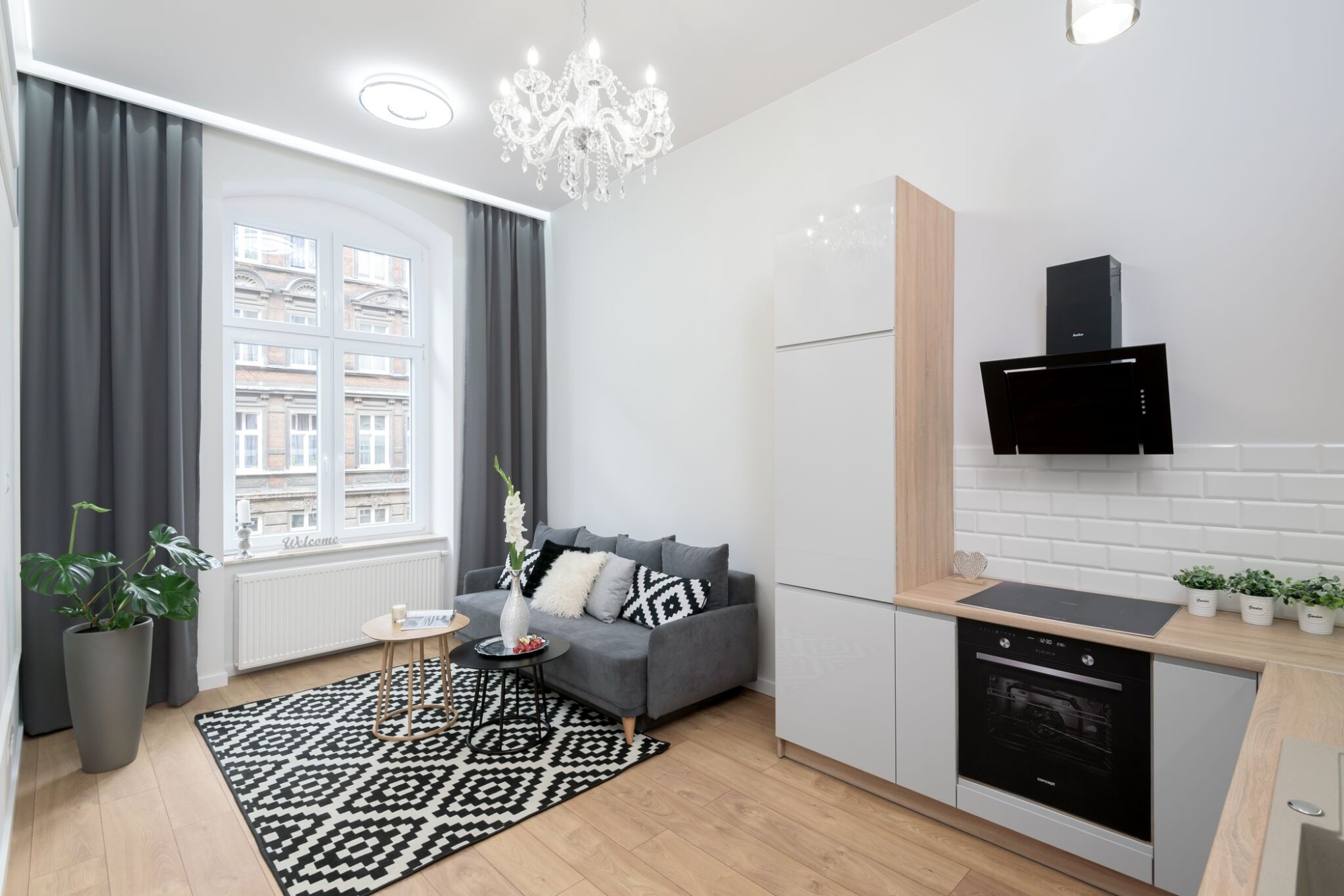 Nowoczesne mieszkanie z szarą sofą, czarno-białym dywanem i kuchnią z białymi szafkami, okapem oraz piekarnikiem w zabudowie.
