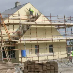 Budowa domu: robotnicy na rusztowaniu podczas nakładania tynku na elewację budynku w trakcie budowy, widoczne materiały budowlane i narzędzia.